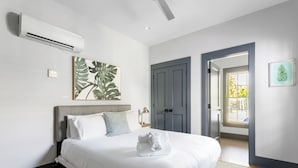 Un lit bien fait, avec du linge de lit blanc et une serviette pliée, une tête de lit grise, une grande œuvre d’art ornée de motifs de feuilles, une porte grise et une fenêtre avec des stores.