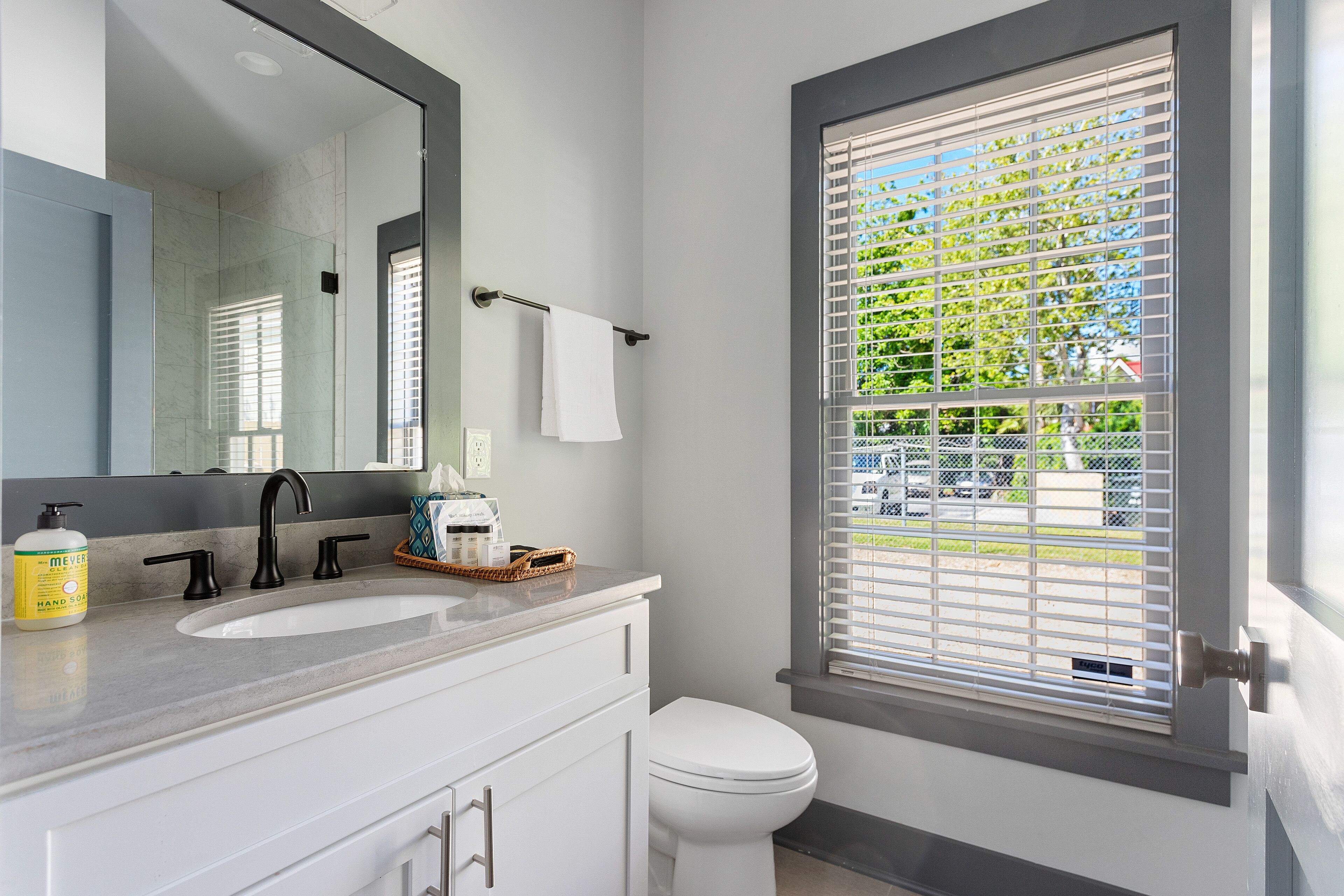 Une salle de bain avec un lavabo blanc, un robinet noir et un grand miroir. Il y a une fenêtre avec des stores et une serviette blanche est accrochée à un porte-serviettes.