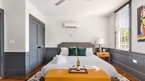 A hotel room with a bed, a ceiling fan, an air conditioning unit, a bedside table with a lamp, a window with blinds, and a painting on the wall. - Only 2 Blocks to King | The Gemstone (Suite B) (Charleston)
