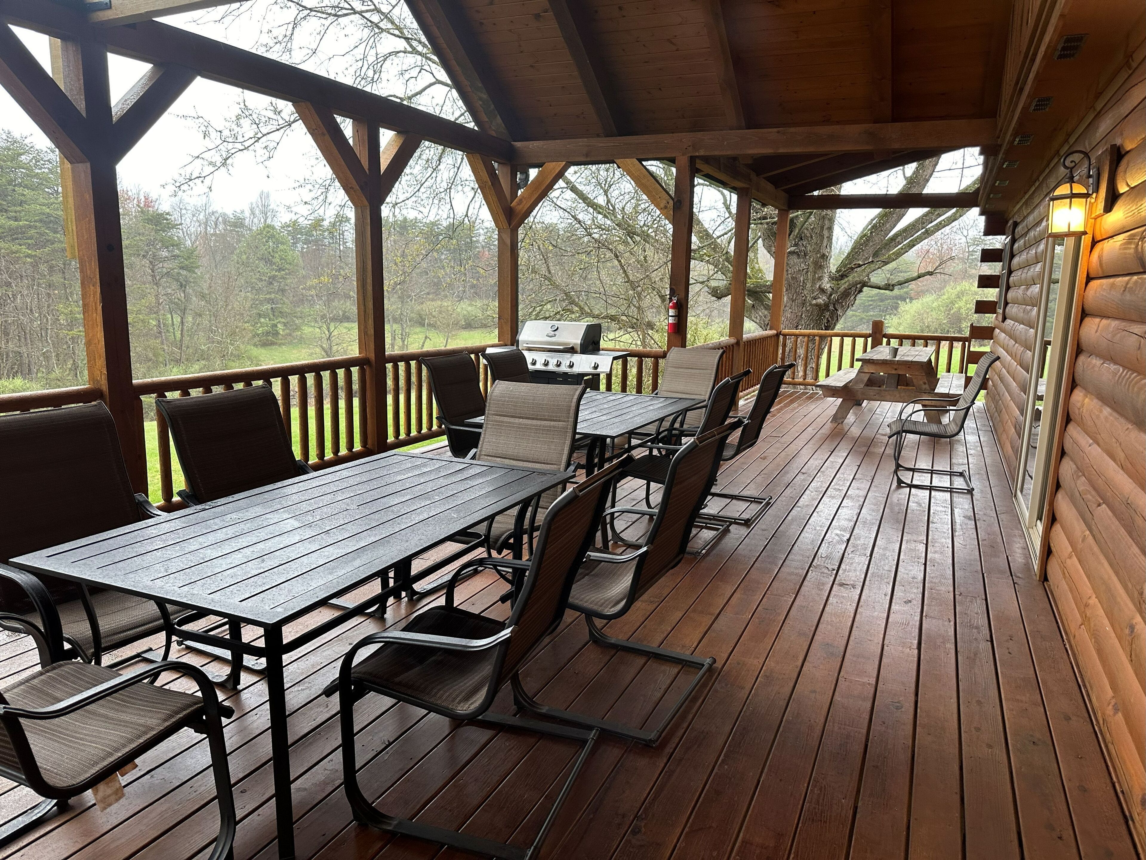 A covered outdoor deck with wooden tables and chairs, a grill, and a view of the surrounding trees.