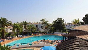A swimming pool surrounded by lounge chairs and umbrellas, with palm trees and buildings in the background.