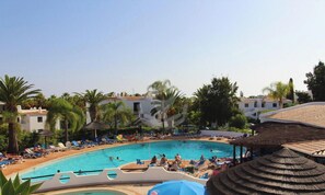 A swimming pool surrounded by lounge chairs and umbrellas, with palm trees and buildings in the background.