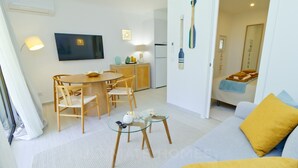 A modern living room with a sofa, a dining table, and chairs. There is a TV mounted on the wall, a small table with a remote control, and a kitchen area visible through an open door.