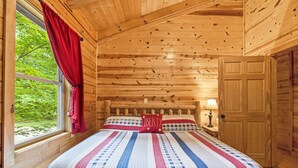 A wooden cabin bedroom with a bed, a window with curtains, and a door. - Freedom Lodge | Sleeps 12 | 4BR + Loft | Hot Tub, Pool Table & Covered Decks (Rockbridge)