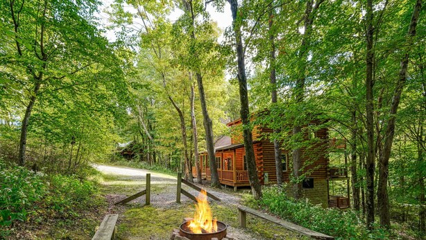 A rustic cabin surrounded by dense forest, a fire pit in the foreground, and a gravel path leading to the cabin. - Freedom Lodge | Sleeps 12 | 4BR + Loft | Hot Tub, Pool Table & Covered Decks (Rockbridge)