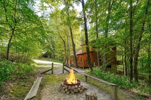 A rustic cabin surrounded by dense forest, a fire pit in the foreground, and a gravel path leading to the cabin. - Freedom Lodge | Sleeps 12 | 4BR + Loft | Hot Tub, Pool Table & Covered Decks (Rockbridge)