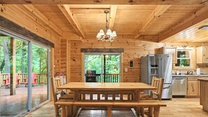 A wooden cabin interior with a dining table, chairs, a kitchen area, and large windows overlooking a forested area. - Freedom Lodge | Sleeps 12 | 4BR + Loft | Hot Tub, Pool Table & Covered Decks (Rockbridge)