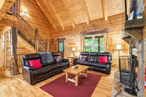 A log cabin interior with two leather sofas, a wooden coffee table, a stone fireplace, and a wooden staircase.