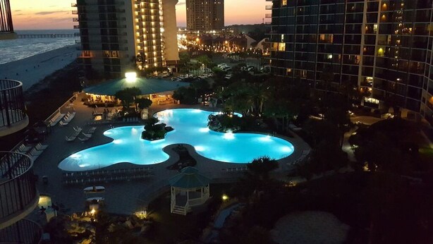 Un área de piscina de hotel con una piscina grande y de forma irregular, rodeada de sillas de descanso y un pabellón.