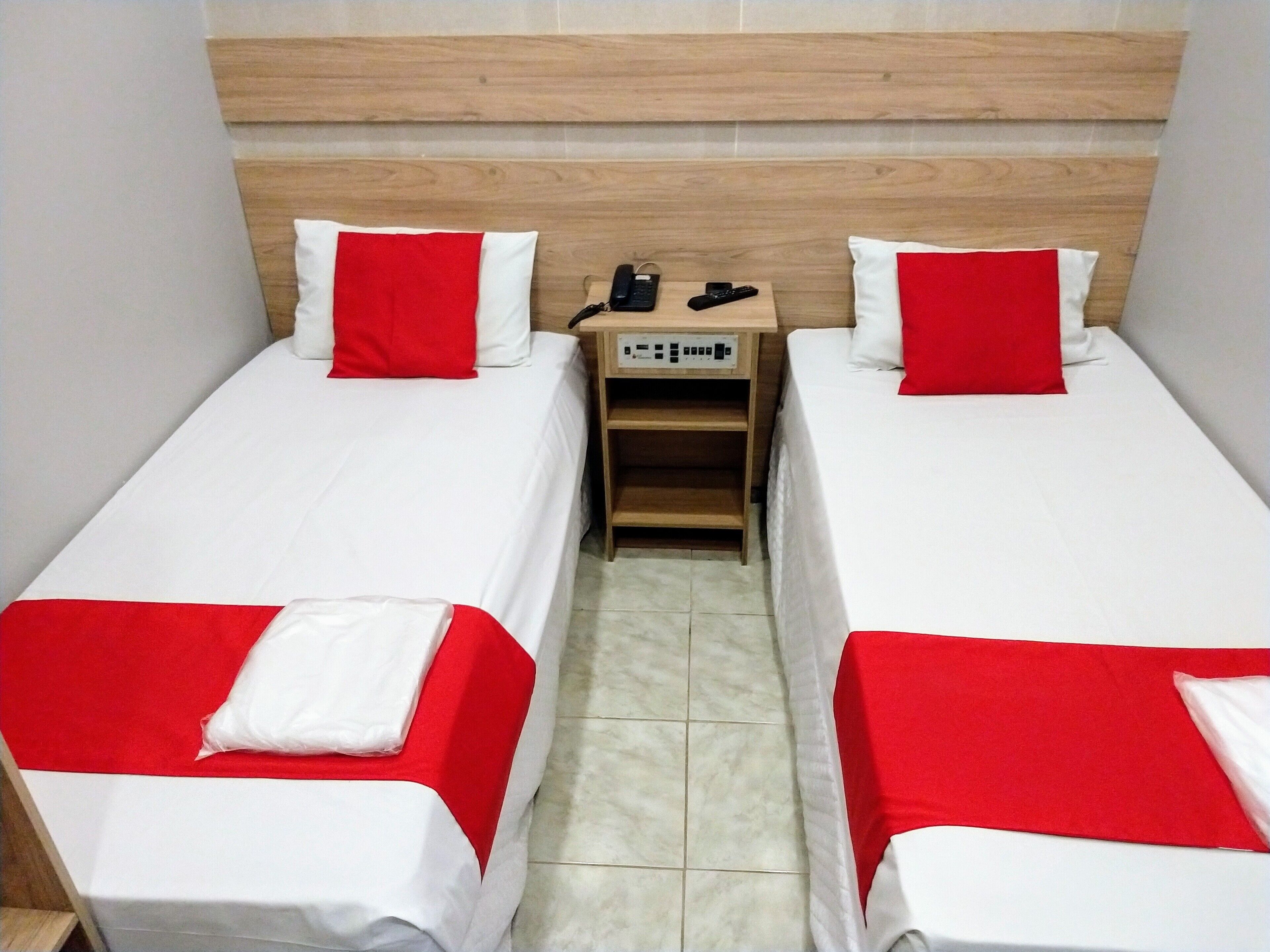 A hotel room with two single beds, each with a red and white bedspread and pillows. There is a wooden nightstand between the beds with a phone and remote controls.