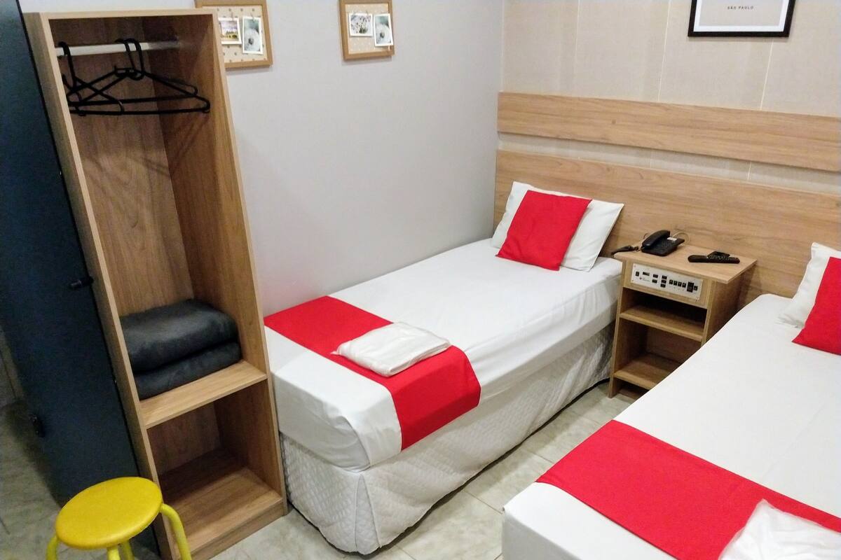 Une chambre d’hôtel avec deux lits, une armoire en bois, un petit tabouret jaune et une table de chevet avec un téléphone et une prise électrique.