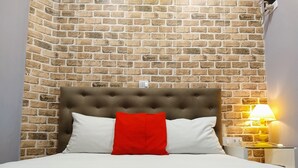 A bed with a tufted headboard, white and red pillows, and a red bedspread against a brick wall backdrop. - Hotel Urban Mooca (São Paulo)