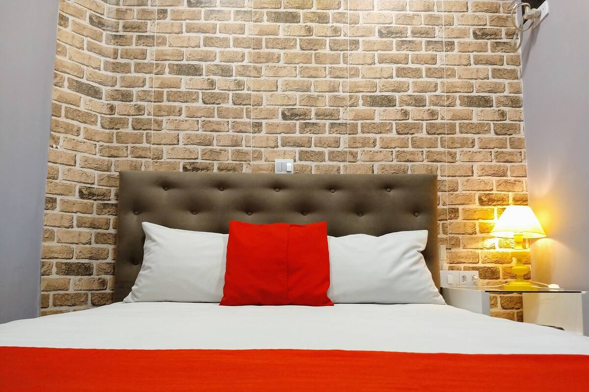 Un lit doté d’une tête de lit capitonnée, de coussins blancs et rouges, et d’une couvre-lit rouge, le tout placé devant un mur en briques.