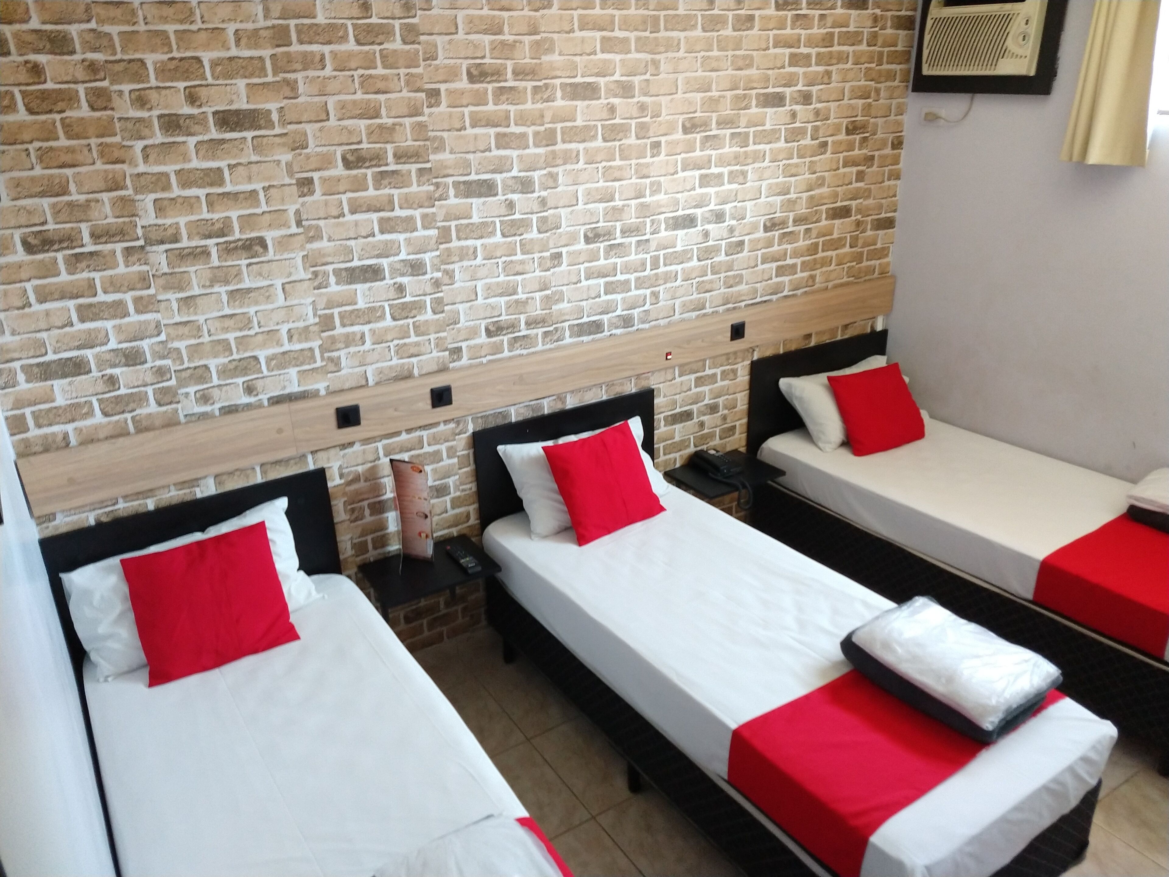 Habitación de hotel con tres camas individuales, cada una con ropa de cama blanca y almohadas rojas. La habitación tiene una pared de ladrillo y un cabecero de madera.