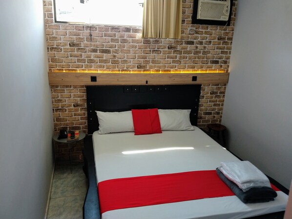 Una cama pequeña y bien hecha con colcha roja y blanca, cabecera negra y repisa de madera arriba.