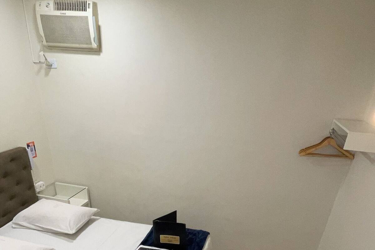 Une petite chambre à coucher, bien rangée, avec un lit, une table de chevet, une lampe suspendue, un climatiseur fixé au mur et une chaise en bois.