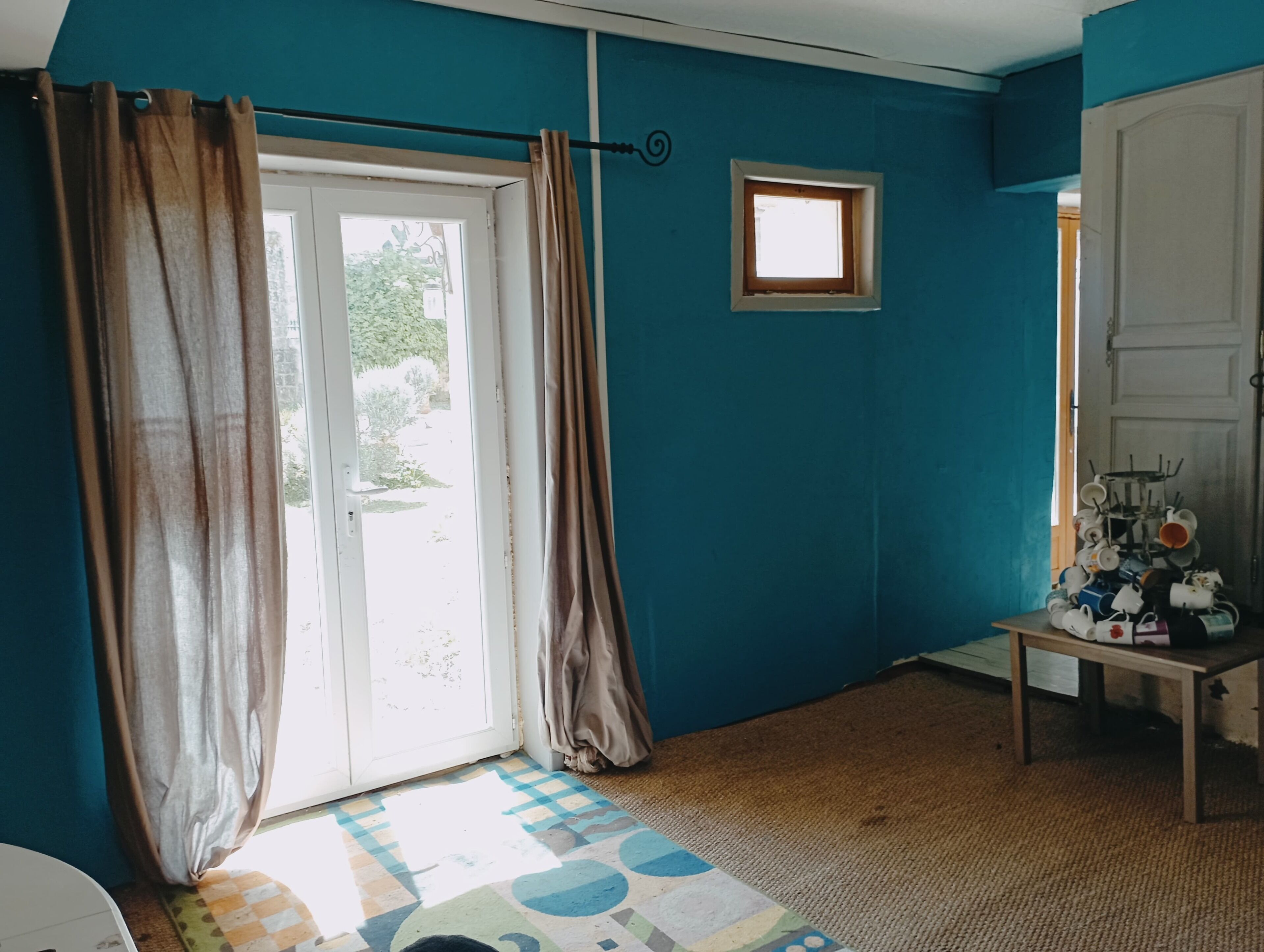 A room with teal walls, a checkered floor, a door with curtains, a small window, and a table with cups.
