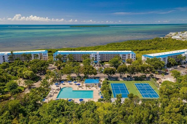 A resort with multiple buildings, a swimming pool, tennis courts, and a view of the ocean.