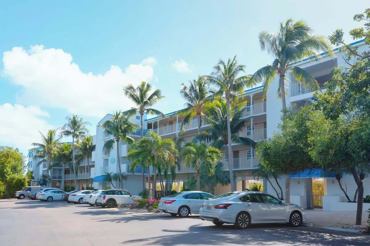 A multi-story hotel with a white exterior and balconies. There are several cars parked in front, and palm trees are visible.