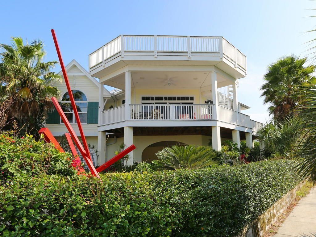 A large, two-story house with a balcony, surrounded by lush greenery and palm trees.