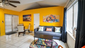 A living room with a gray sofa, a coffee table, and a colorful rug. There is a ceiling fan, a window with blinds, and a door with a doormat.