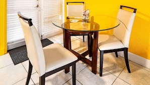 A dining area with a glass table and four chairs, a painting on the wall, and a door with blinds.