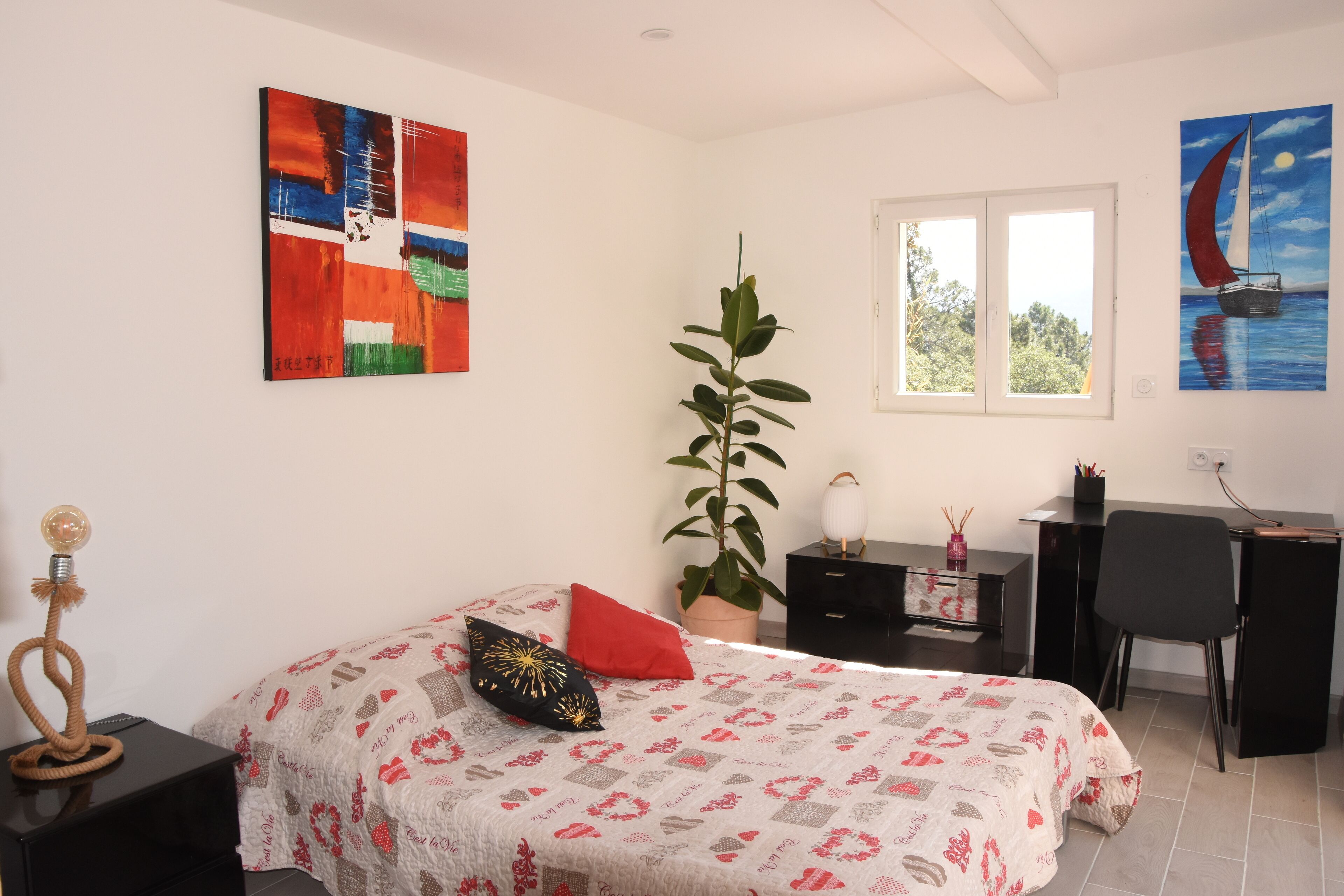 A bedroom with a bed, a desk, a chair, a potted plant, and artwork on the walls.