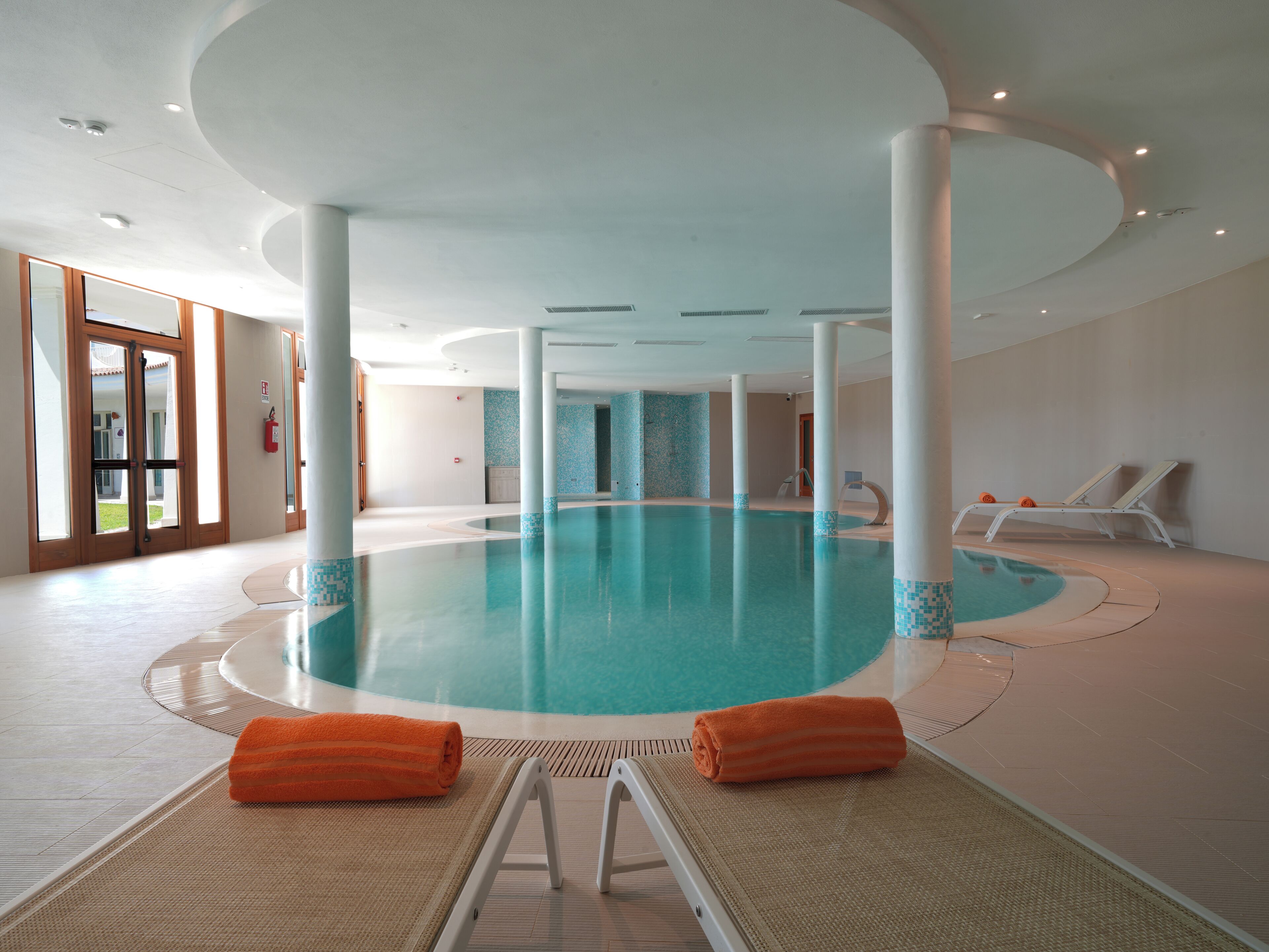Une grande piscine intérieure à forme circulaire, entourée de chaises longues et offrant une vue dégagée sur l’extérieur.