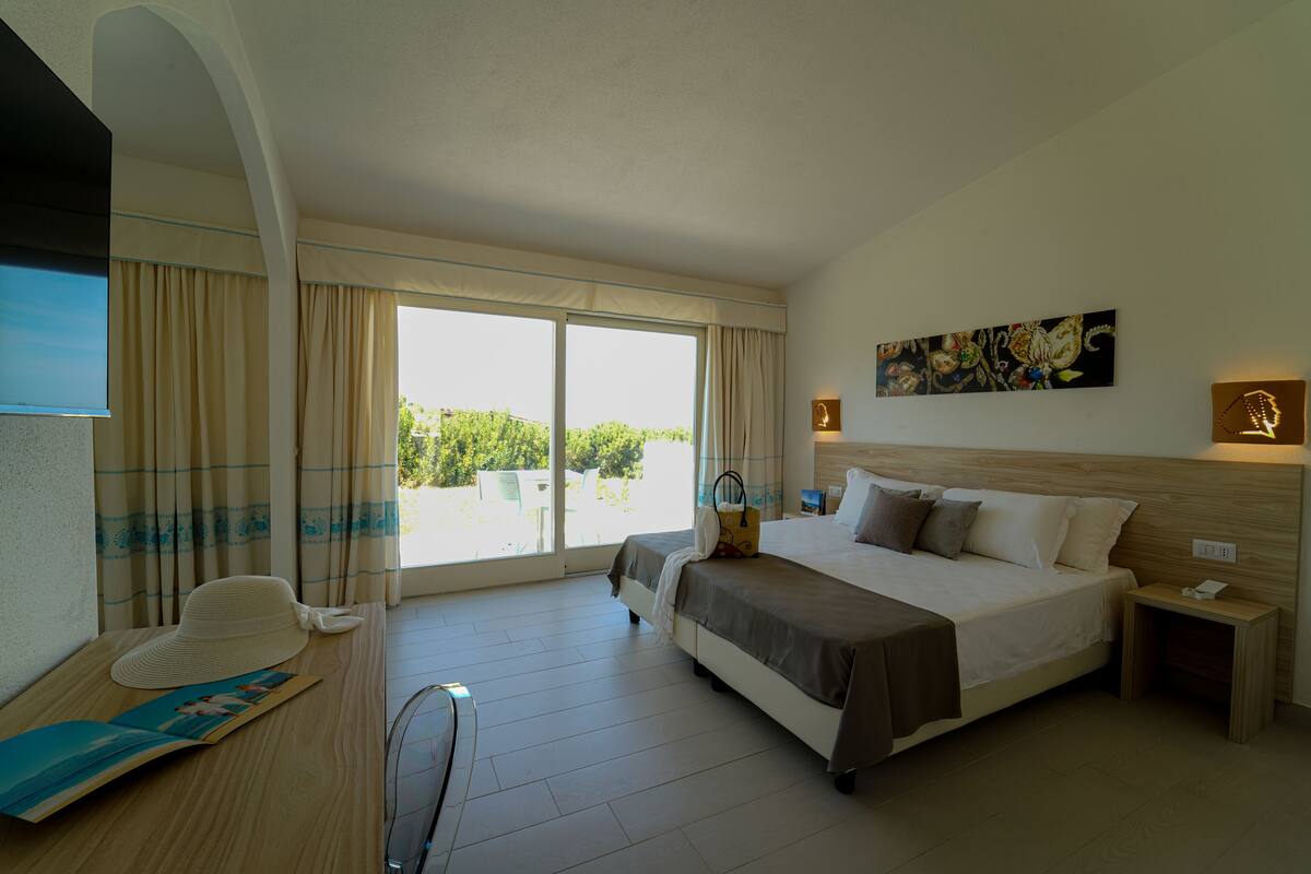 A hotel room with a large bed, a desk with a chair, a window with curtains, and a painting on the wall.