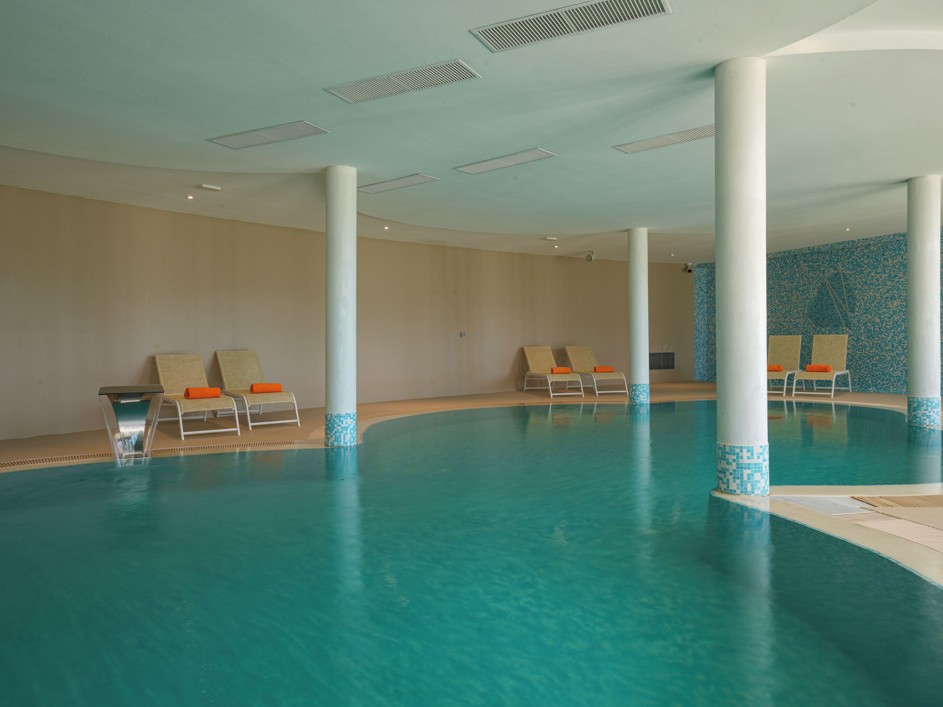 A large indoor swimming pool with clear blue water, surrounded by white columns and lounge chairs with orange cushions.