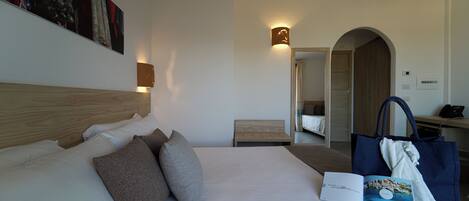 A hotel room with a large bed, a bedside table, a reading lamp, a wall-mounted artwork, and a door leading to another room.