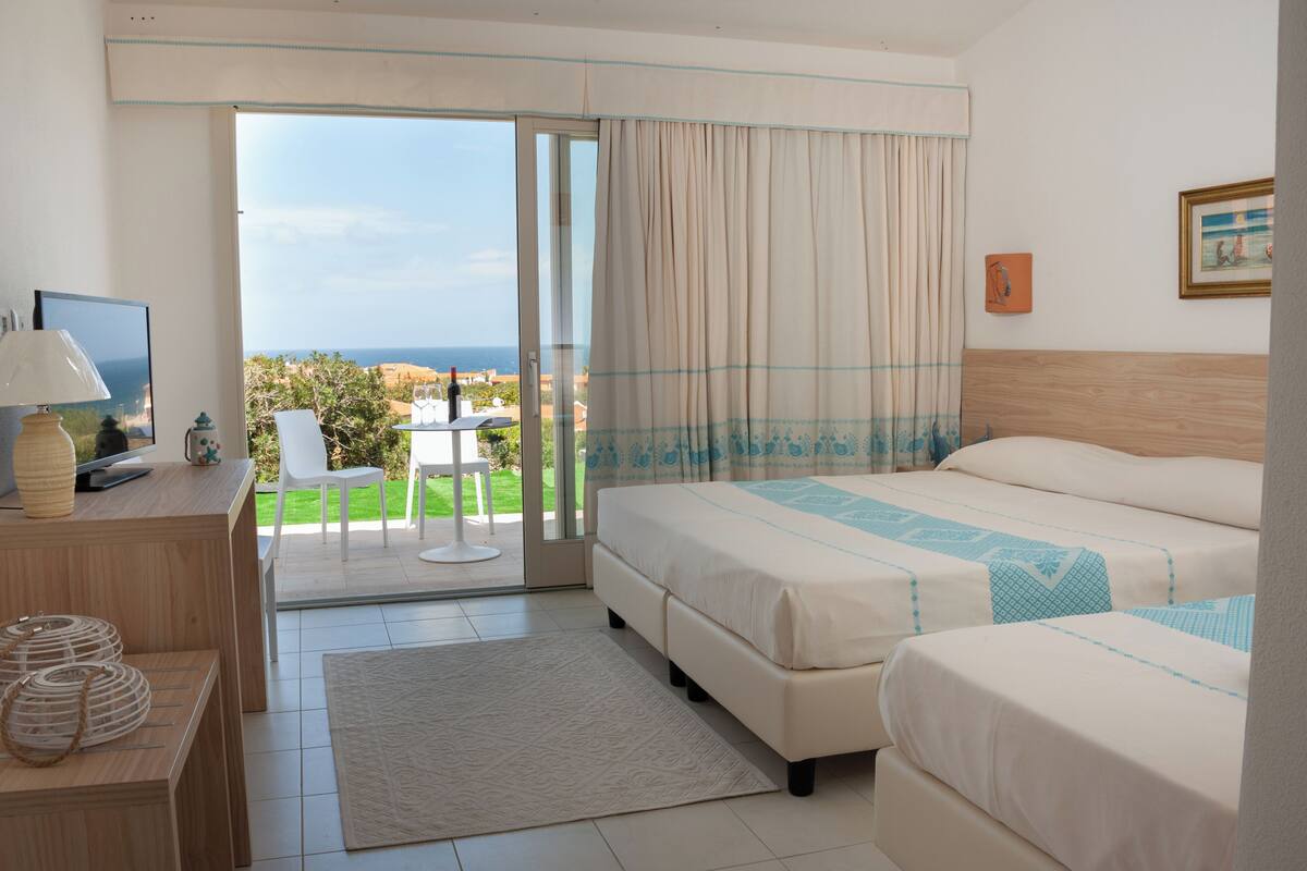 A hotel room with a bed, a desk with a lamp, a TV, and a view of the sea through a sliding door.