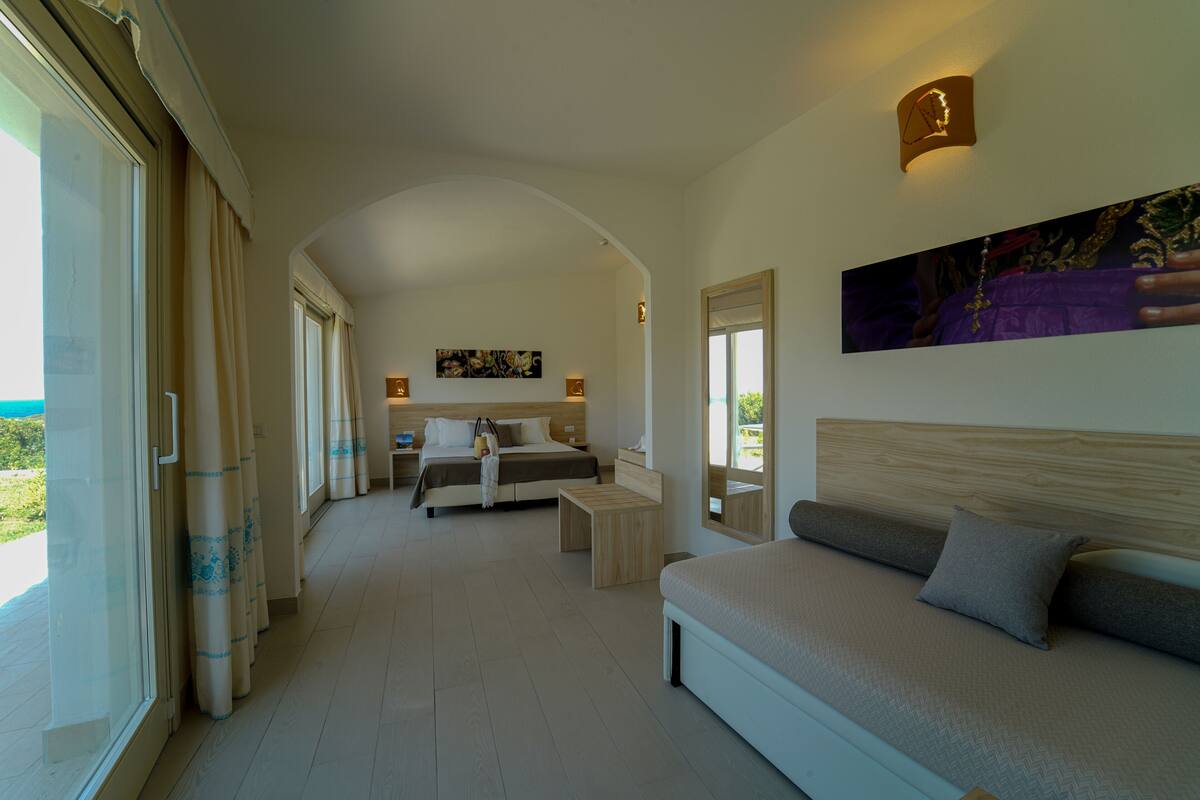 A bedroom with a large bed, a sofa, and a desk. There is a window with curtains, a painting on the wall, and a view of the outdoors.