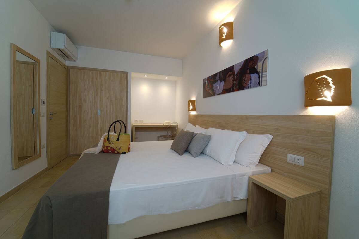 A hotel room with a bed, wooden wardrobe, bedside table, and a wall-mounted picture.