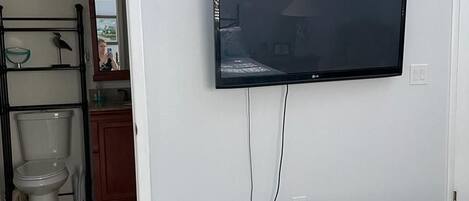 A bedroom with a bed, a television mounted on the wall, a bathroom with a toilet and sink, and a mirror.