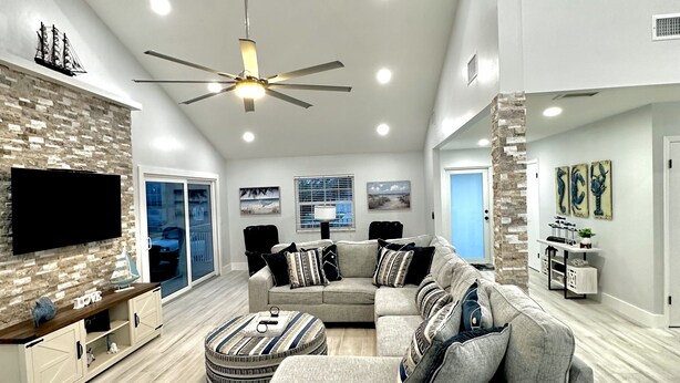A spacious living room with a large gray sectional sofa, a round ottoman, a flat-screen TV mounted on a brick wall, and a ceiling fan. - Breath taking canal, pool home with golf access. 15 minutes from beaches. (Port Charlotte)