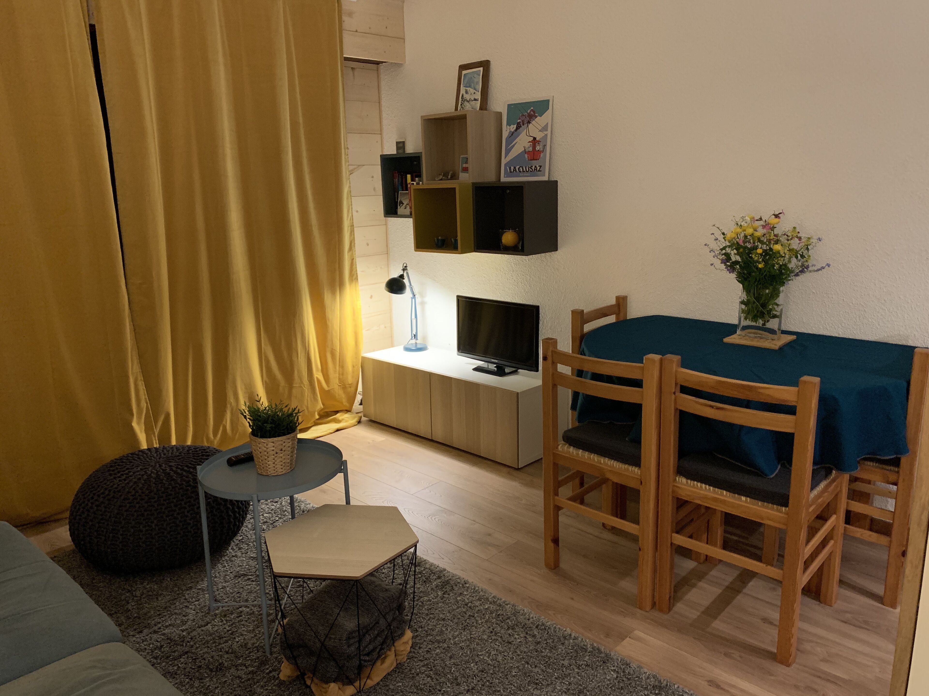 A cozy living room with a sofa, a small round table, a wooden dining table with chairs, a TV on a stand, and a bookshelf with a picture frame.