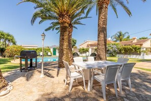 Um pátio externo com mesa e cadeiras, uma churrasqueira e uma piscina ao fundo.