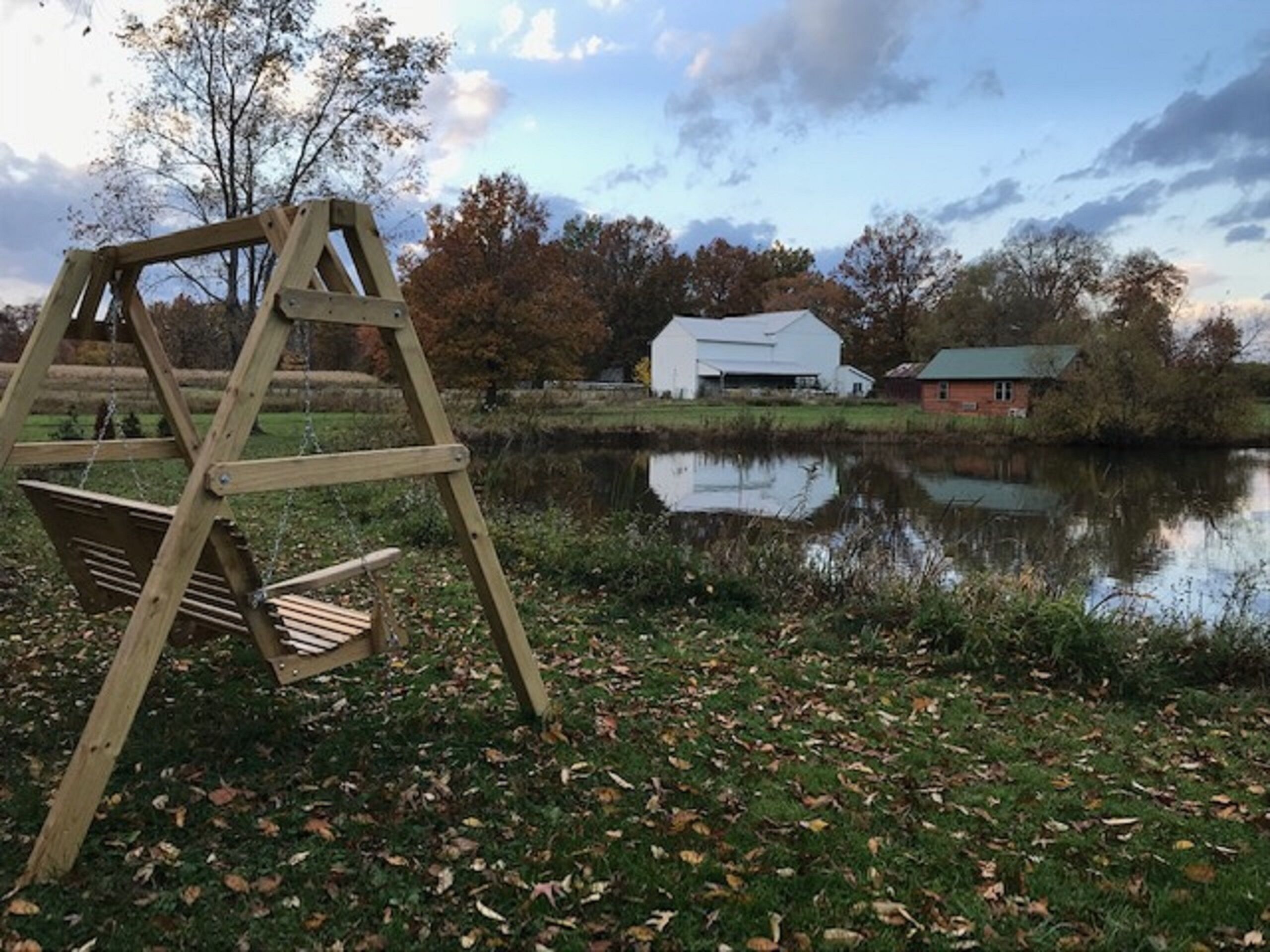 Un columpio de madera cerca de un lago tranquilo, con un granero y una casa al fondo.