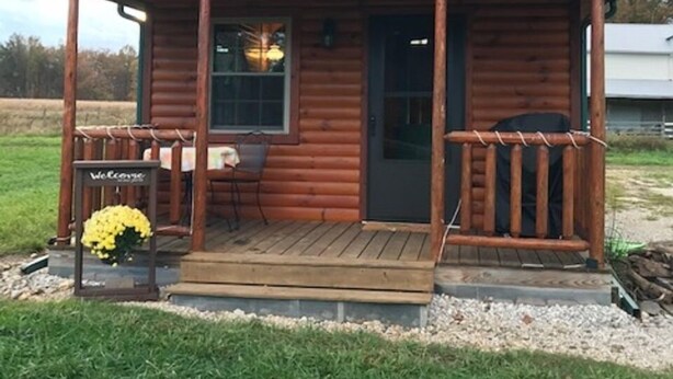 Une cabane en rondins avec un porche, une pancarte de bienvenue et une fenêtre.