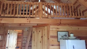 L’intérieur d’un chalet en bois comprend une chambre à coucher située en mezzanine, un coin cuisine avec un réfrigérateur et une porte donnant sur une autre pièce.