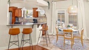 Una cocina moderna con armarios de madera, una isla central y taburetes de bar. Hay un comedor con una mesa redonda y sillas, y una ventana con cortinas.