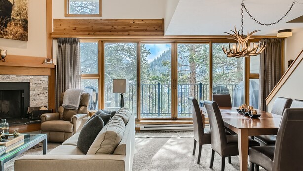 A spacious living room with a fireplace, leather sofa, dining table, and large windows offering a view of trees and mountains. - Newly Updated Condo, Pool & Hot Tub, Shuttle to Slopes, Large Private Patio (Keystone)