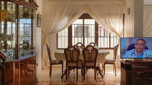 A dining area with a glass cabinet, a round table with chairs, and a television displaying a person.