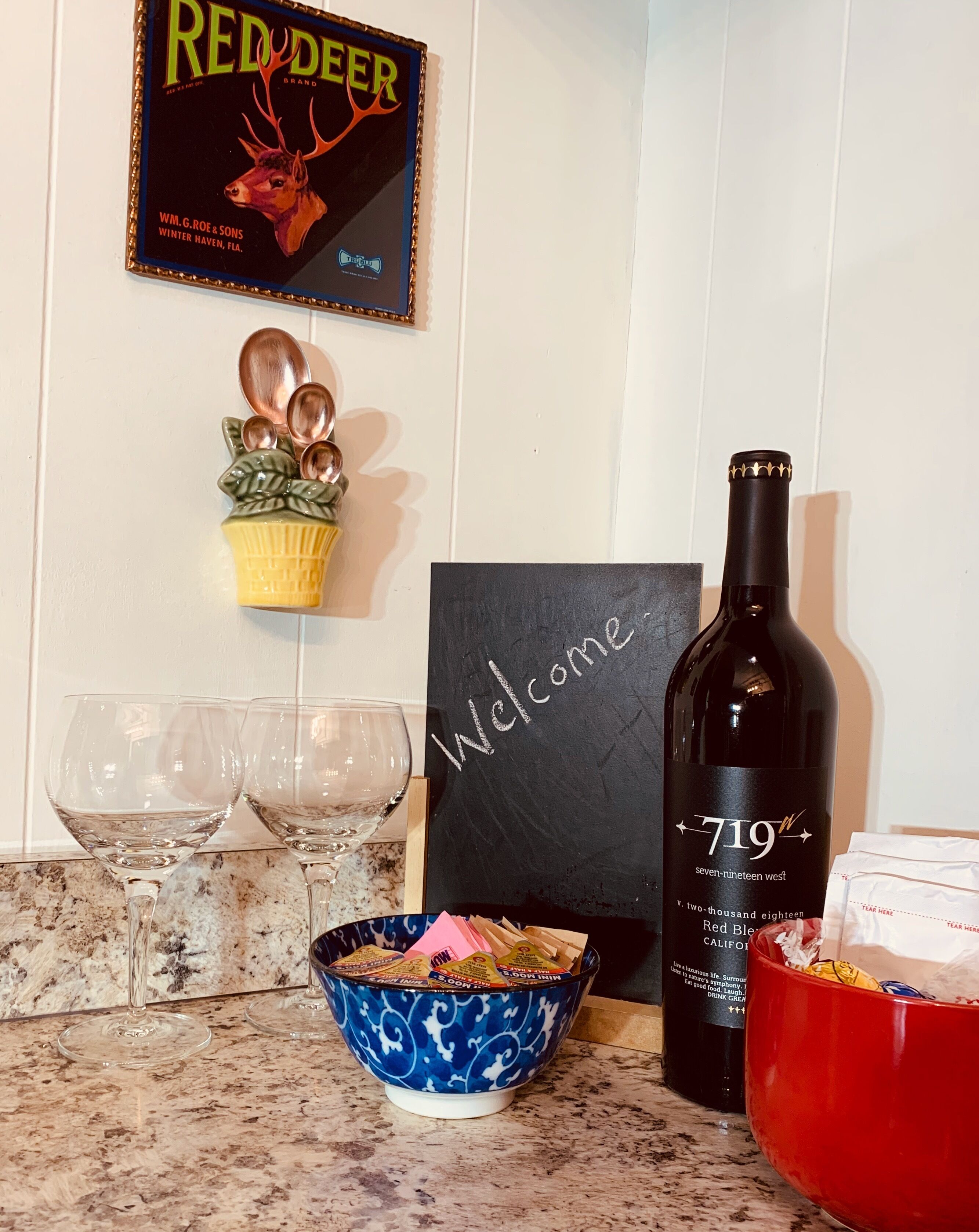 A wine bottle, two wine glasses, a welcome sign, a decorative bowl, and a framed sign on a kitchen counter.