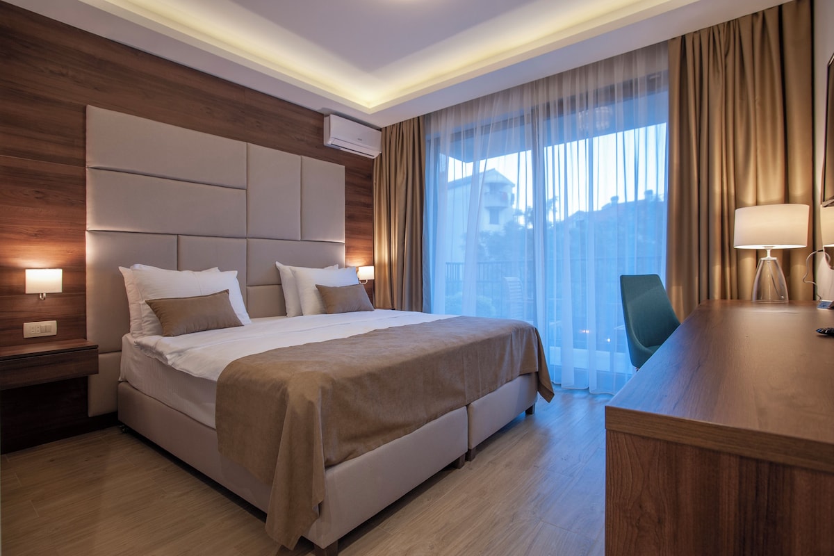 Habitación de hotel con una cama grande, un escritorio, una silla y un ventanal con cortinas.