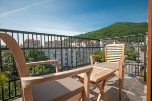 A balcony with two wooden chairs and a table, overlooking a cityscape with buildings and a hill.