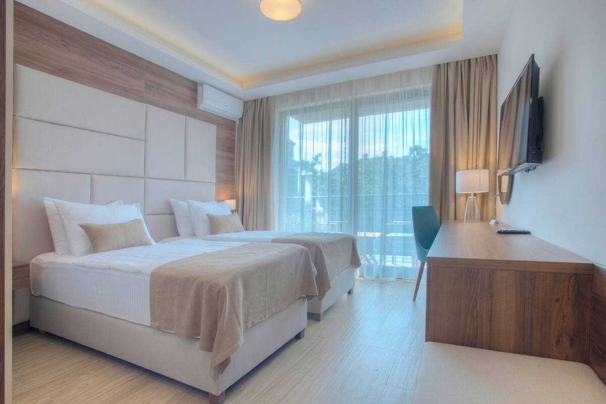 A hotel room with a large bed, a desk, a chair, a TV, and a balcony with curtains.