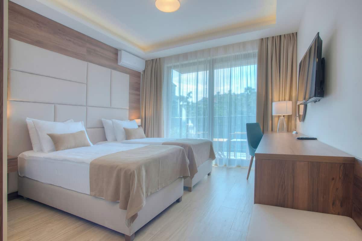 A hotel room with a large bed, a desk, a chair, a TV, and a balcony with curtains.