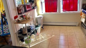 A kitchen with a window, a shelf with kitchen items, a trash bin, and a tiled floor.